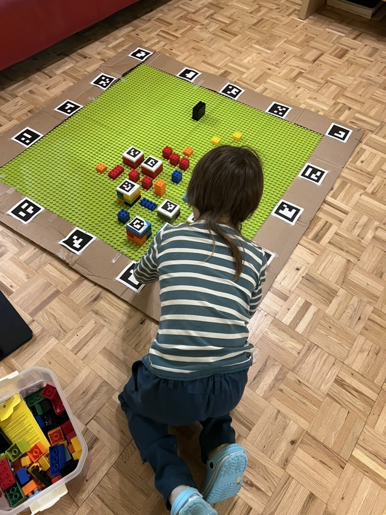 My kiddo kneels at the green board, building; the board sits in a cardboard frame lined with printed black-and-white cards