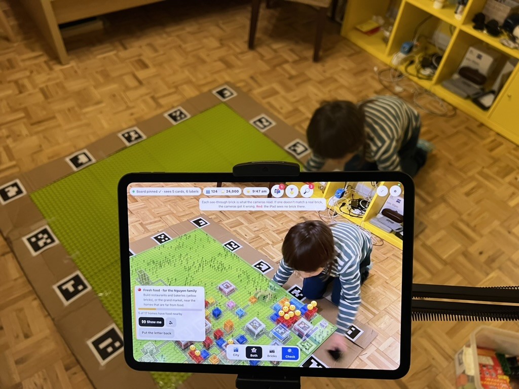 The iPad on its stand in front of the board, in Both mode: see-through colored bricks sit on the real board, with worried faces over some homes; my kiddo builds in the background