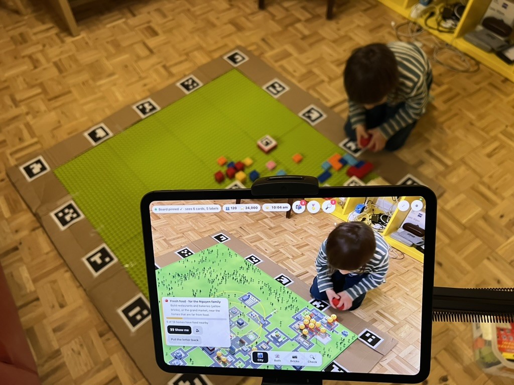 The iPad on its stand in City mode: the bricks have become a town with roads, buildings and trees, while my kiddo holds a red brick in the background