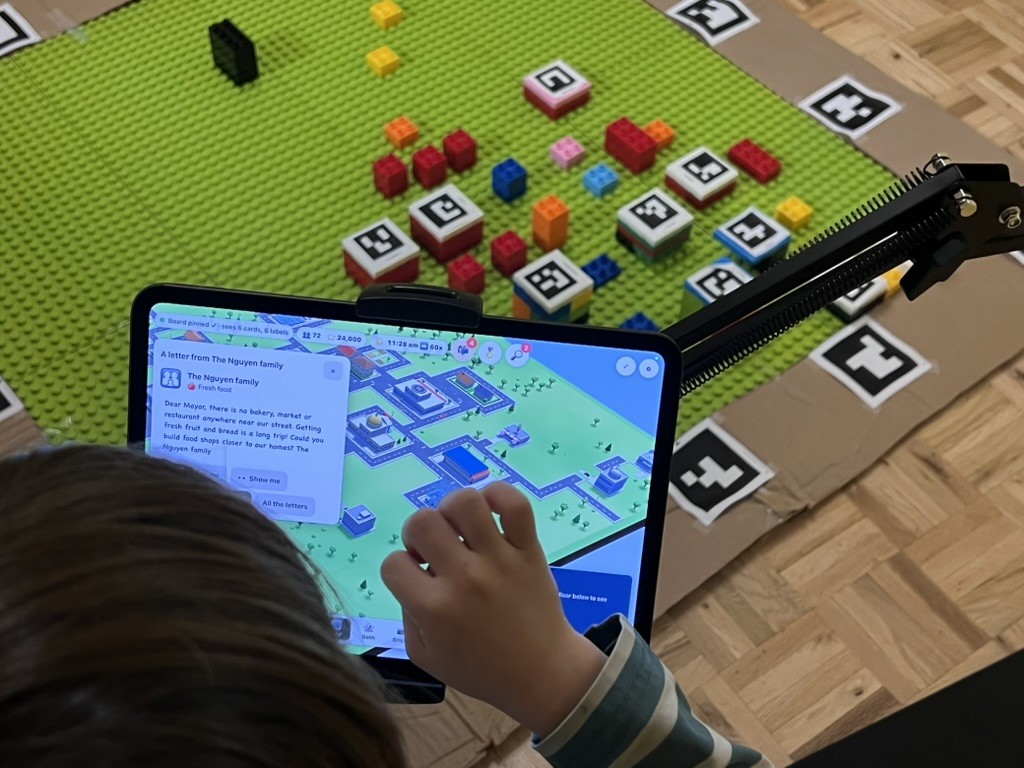 My kiddo touches the iPad screen, where a letter from the Nguyen family is open over the 3D city; the real bricks are on the board behind the iPad
